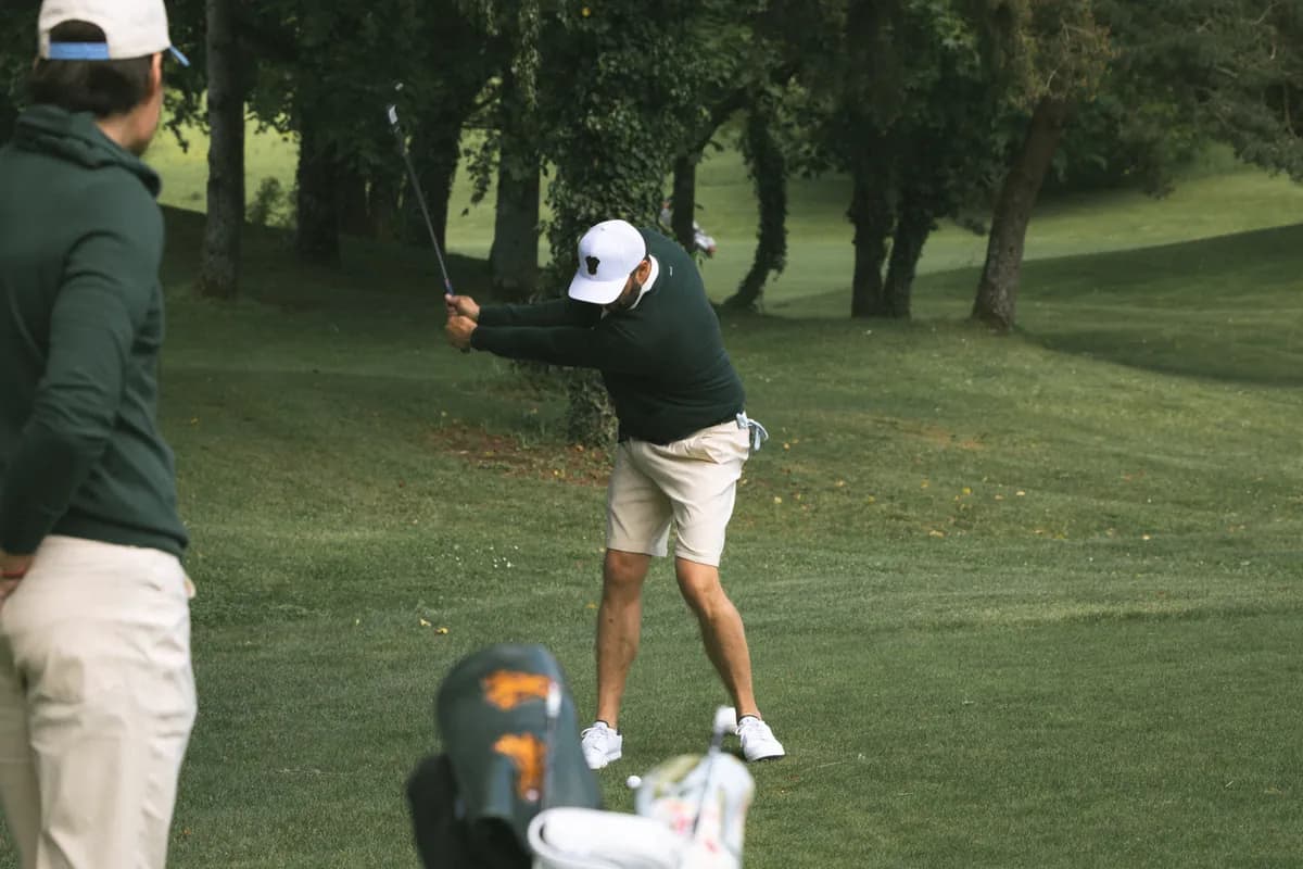 Golfspiller i grøn sweater, der afslutter sit sving, med en anden spiller i forgrunden